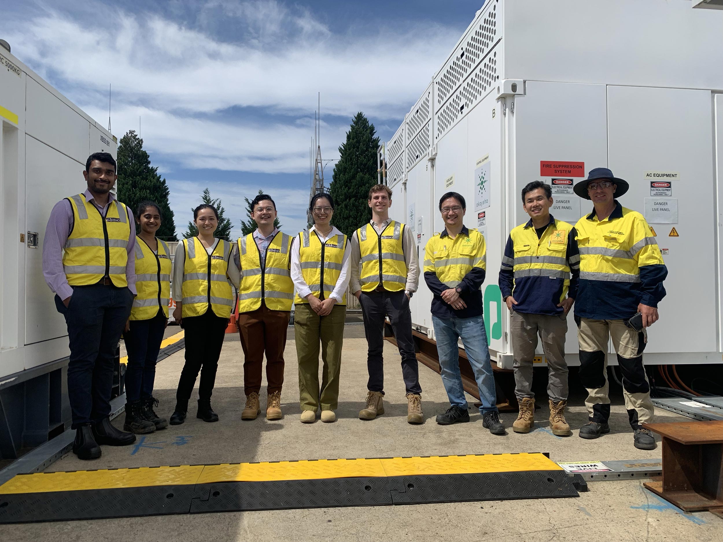 Nine men and women of mixed ethnicity wearing yellow safety vests standing in front of a large BESS array