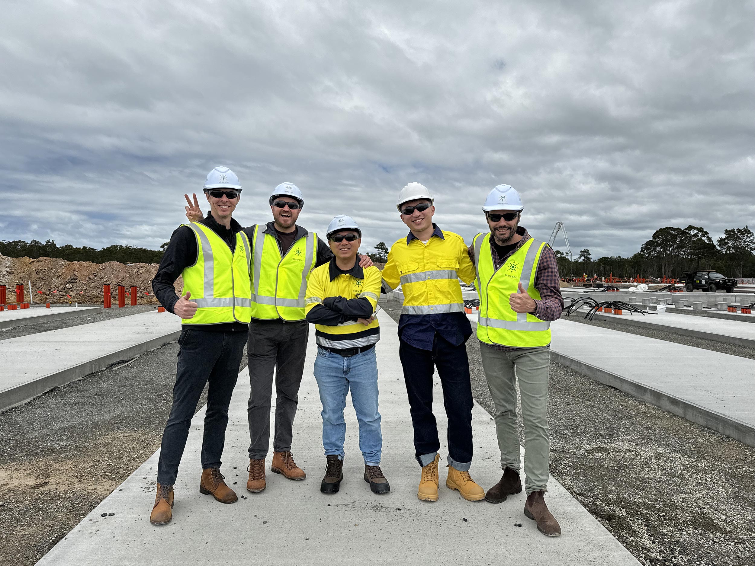 Five mean wearing PPE standing in front of a BESS project under construction smiling at the camera. Two men are displaying a thumbs up gesture and another is showing a peace sign.