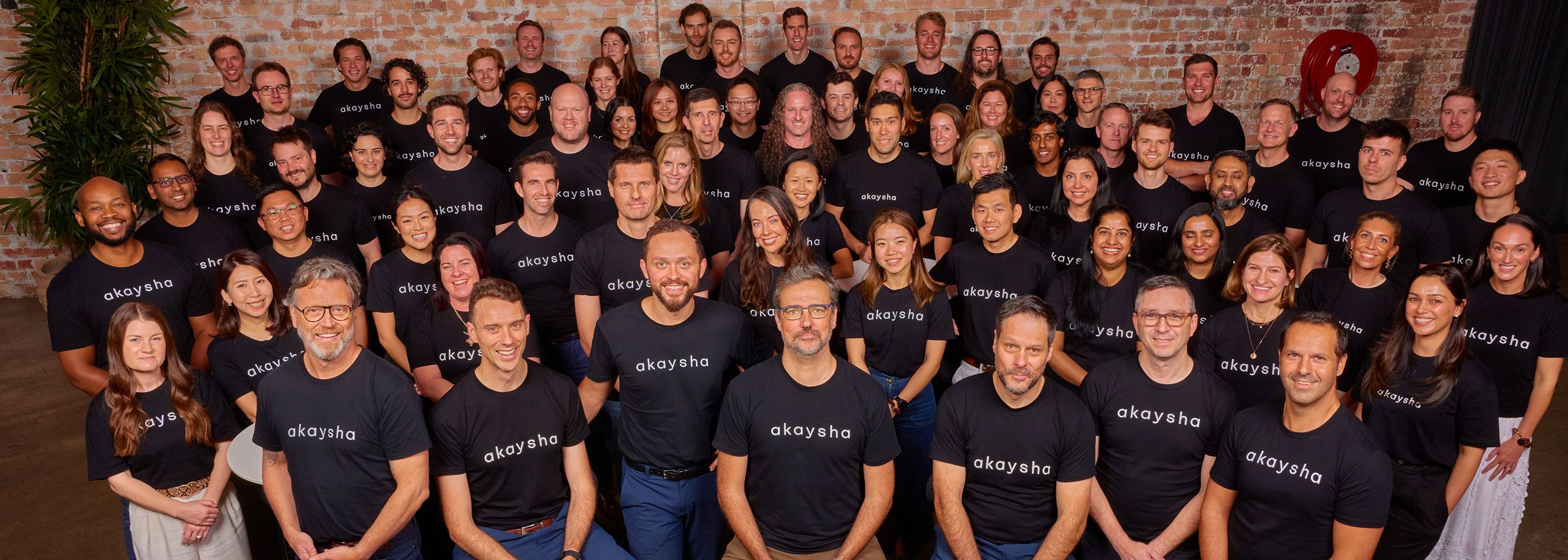 A wide view of the entire Akaysha team posing in front of a brick wall inside an office