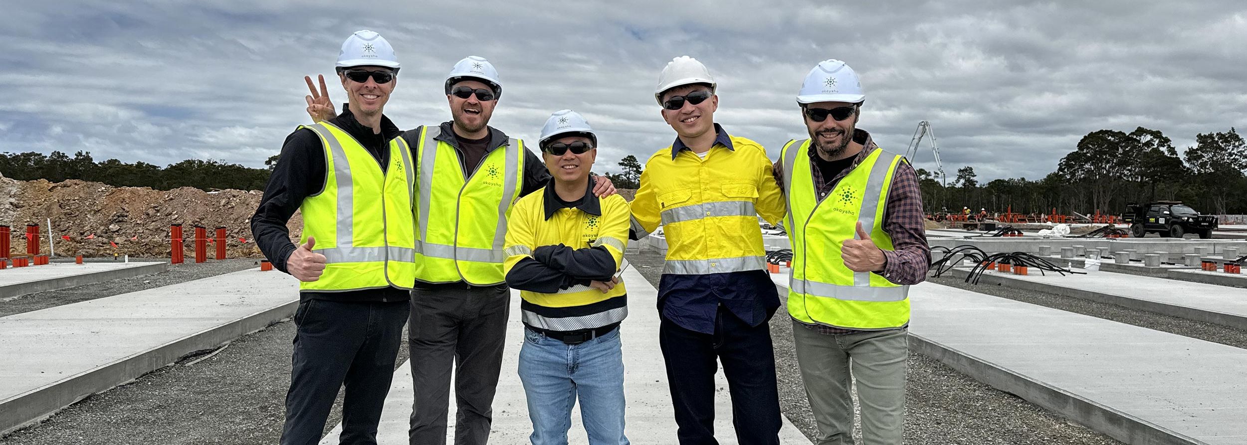 Five mean wearing PPE standing in front of a BESS project under construction smiling at the camera. Two men are displaying a thumbs up gesture and another is showing a peace sign.