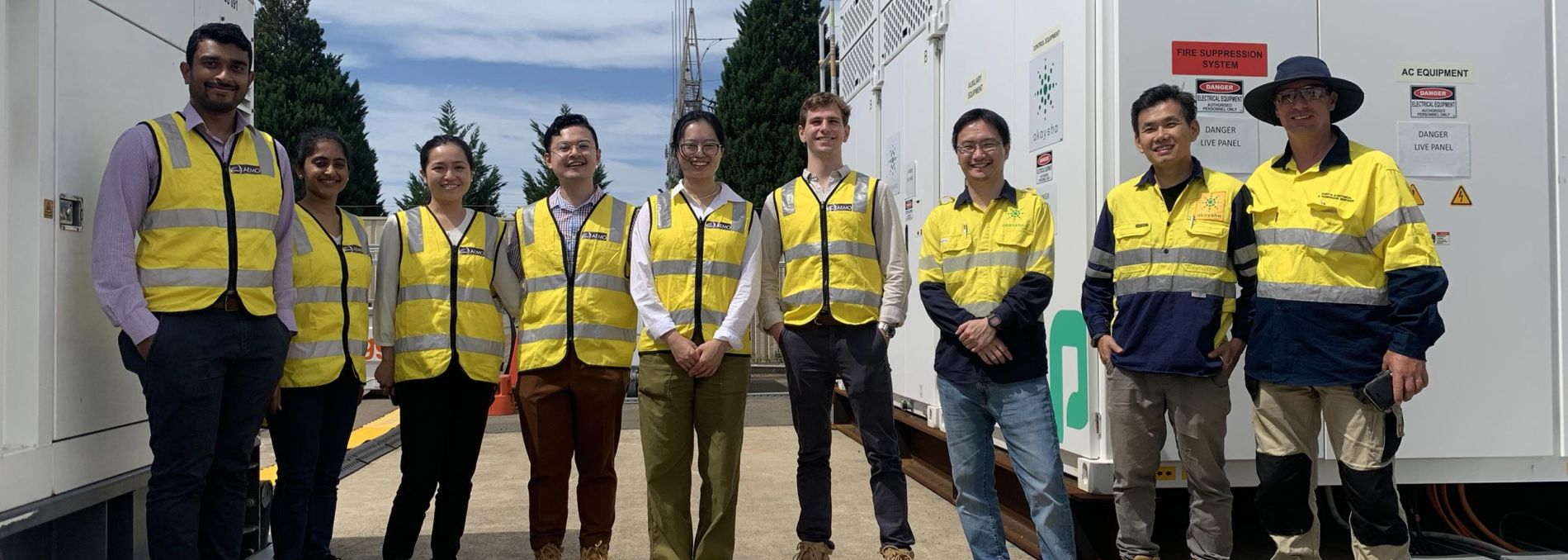 Nine men and women of mixed ethnicity wearing yellow safety vests standing in front of a large BESS array