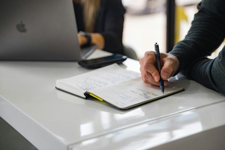 A cropped image of a man writing in a diary at a desk with a woman working on a laptop sitting next to him