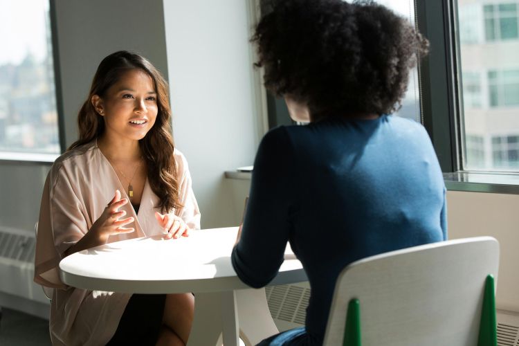 A young asian woman sitting at a small white table talking to a young black woman who has her back to the camera